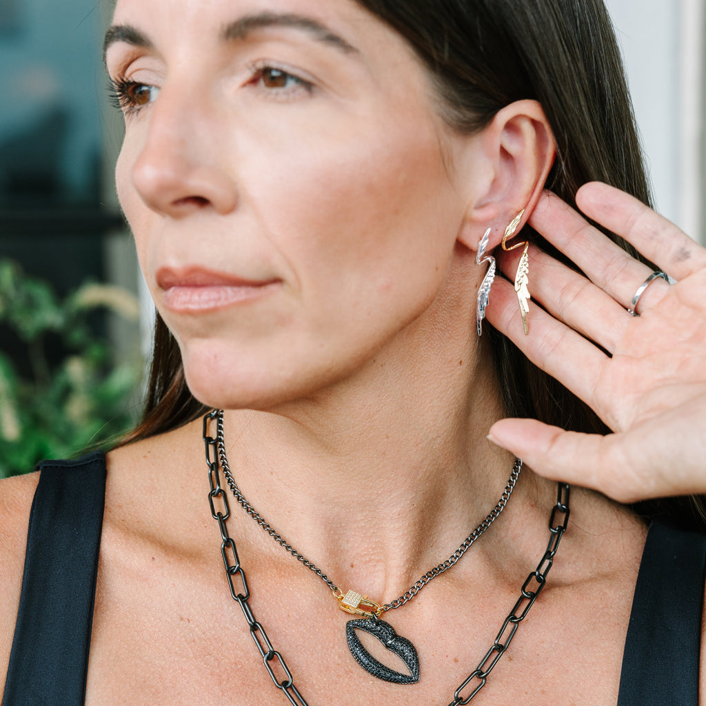 A close-up lifestyle shot of a woman wearing layered black and gold necklaces, including a chain necklace with a lip-shaped pendant and a drop pendant, along with hoop earrings.