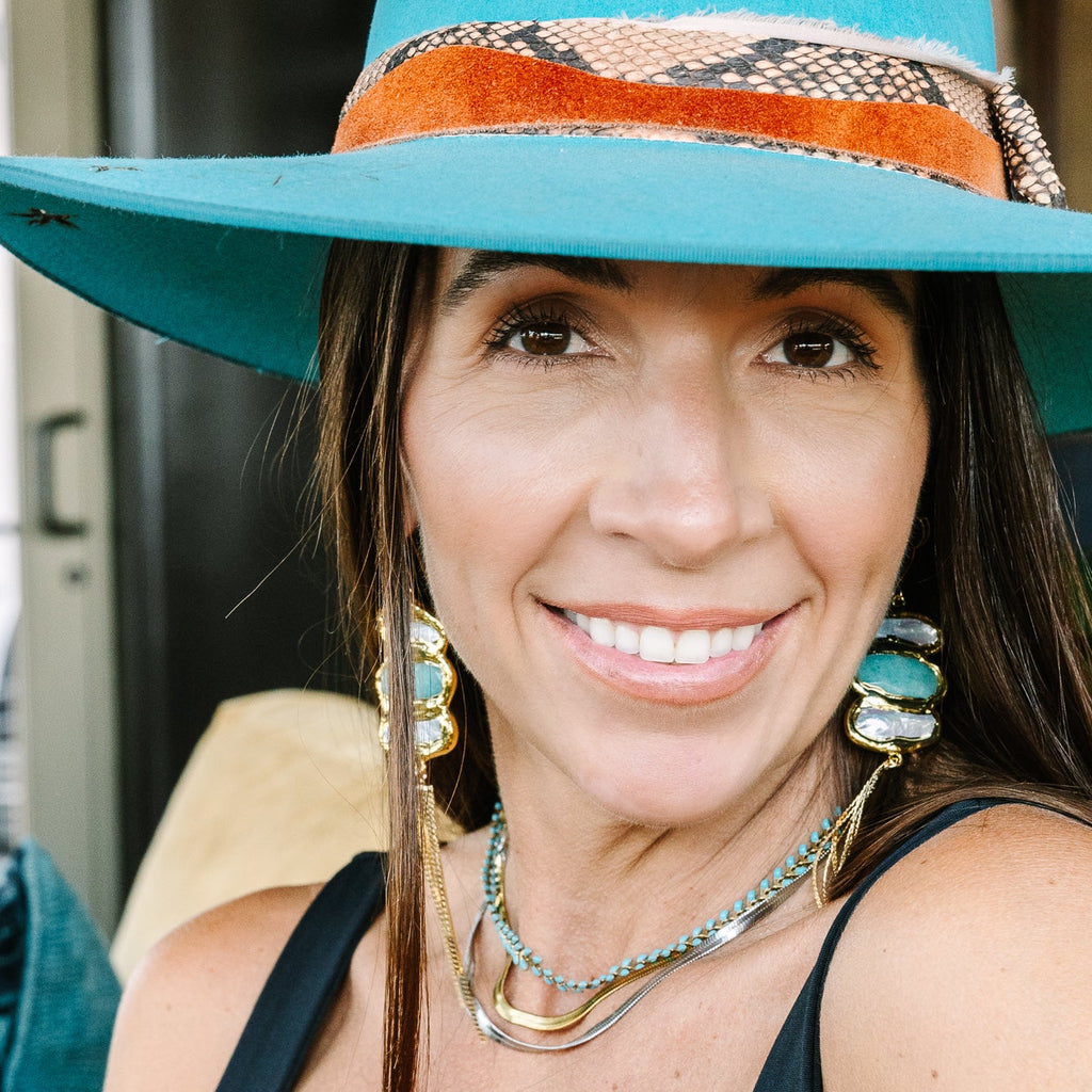 A smiling woman wearing a wide-brim turquoise hat with a patterned orange and brown band, layered silver necklaces, and large turquoise earrings, photographed in a close-up portrait.