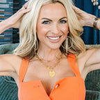A smiling blonde woman posing in a bright orange sleeveless top, wearing gold necklaces and hoop earrings, seated on a green velvet chair in front of patterned blue curtains.