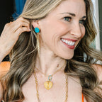 A smiling woman with long wavy brown hair wearing a bright orange top, gold necklaces, and a turquoise earring, shown in a close-up portrait.