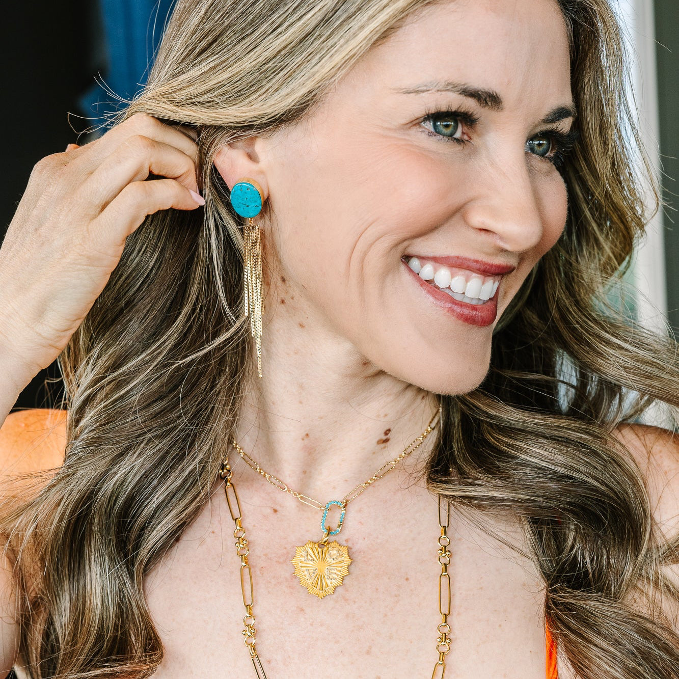 A smiling woman with long wavy brown hair wearing a bright orange top, gold necklaces, and a turquoise earring, shown in a close-up portrait.