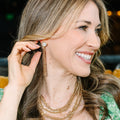 A woman with long wavy brown hair smiling and looking to the side, wearing layered gold necklaces and a long dangling gold earring with a pearl-like bead.