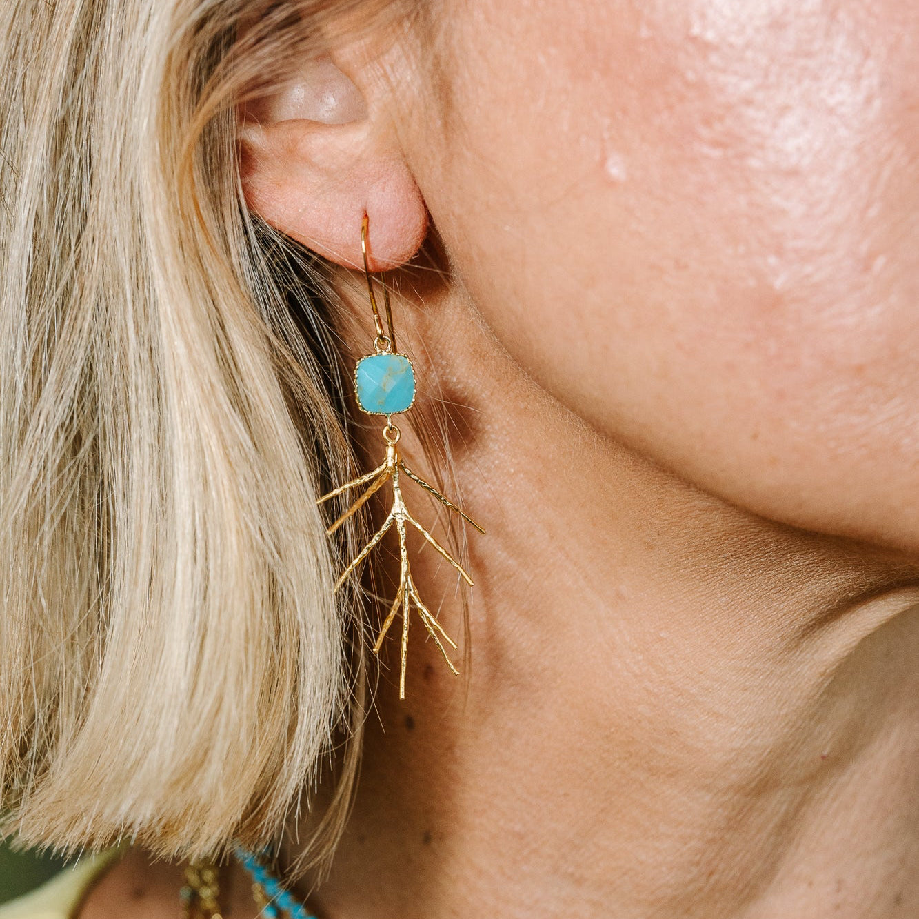 Close-up of a model wearing a gold leaf dangle earring with a round turquoise blue stone, along with layered gold and turquoise necklaces.