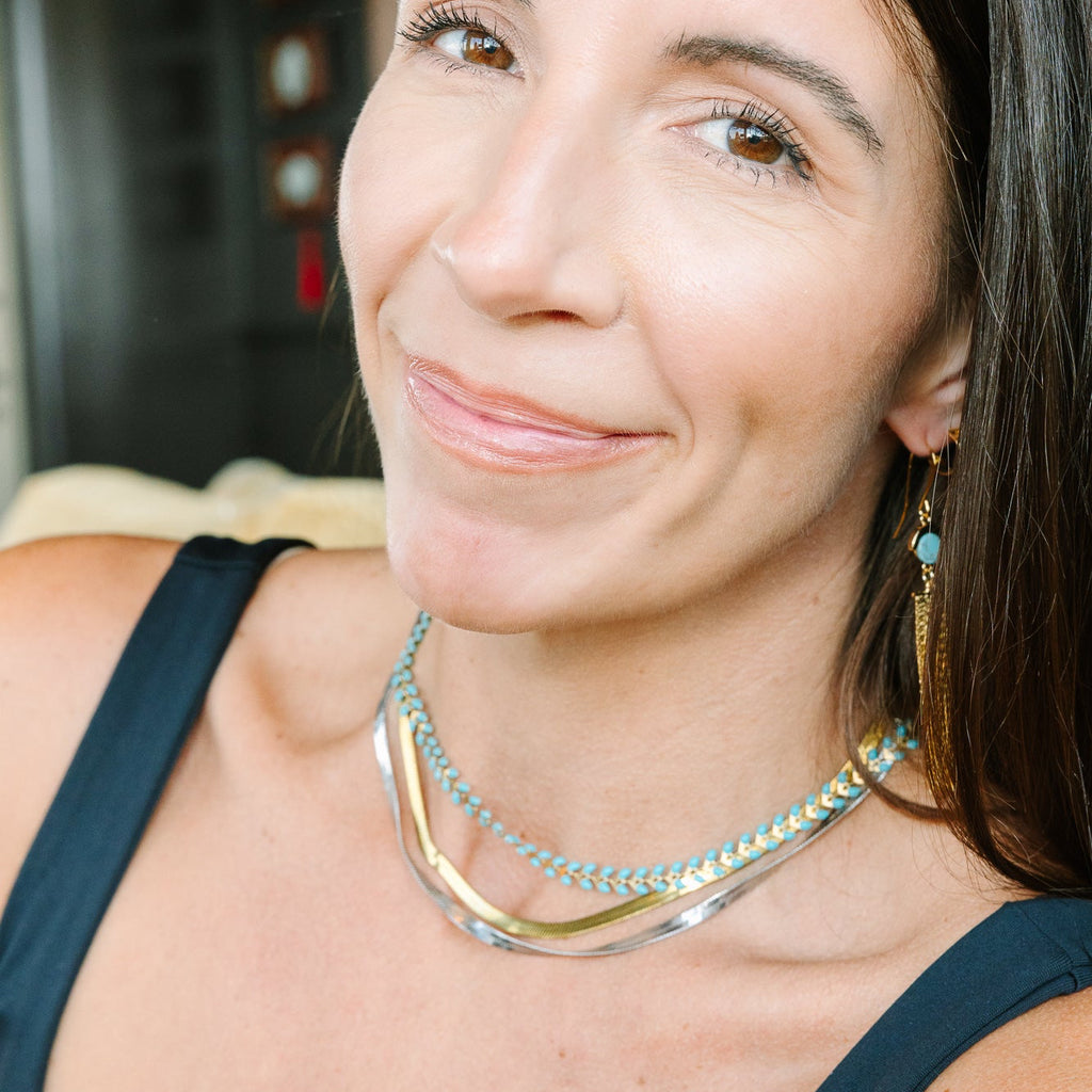 A close-up portrait of a woman wearing a layered necklace with gold and turquoise blue chains, along with matching drop earrings, set against a softly blurred indoor background.