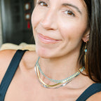 A close-up portrait of a woman wearing a layered necklace with gold and turquoise blue chains, along with matching drop earrings, set against a softly blurred indoor background.