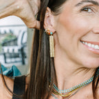 A close-up lifestyle shot of a woman wearing a gold huggie hoop earring with a dangling gold tassel and a small turquoise accent, along with layered gold and turquoise necklaces.