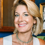 A smiling blonde woman wearing layered gold necklaces, including a horseshoe pendant, and large hoop earrings, wearing a white sleeveless top.