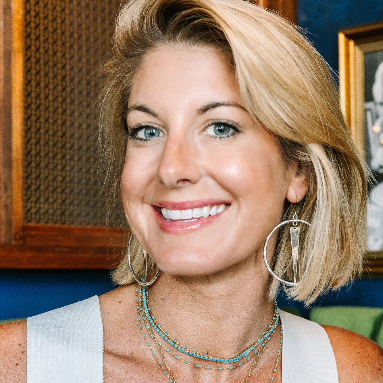 A smiling blonde woman wearing layered gold necklaces, including a horseshoe pendant, and large hoop earrings, wearing a white sleeveless top.