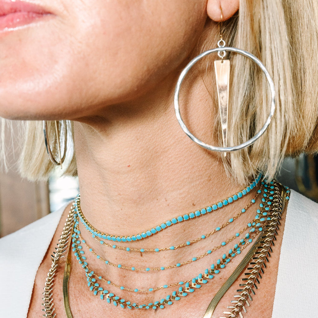 Close-up of a woman wearing layered necklaces, including a turquoise beaded chain, a gold snake chain, and a gold feather-style chain, along with a large silver hoop earring.