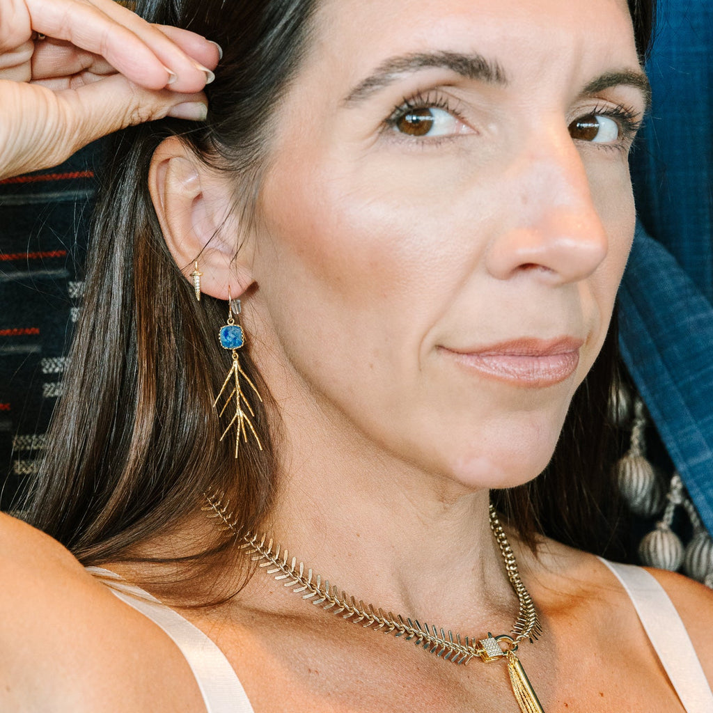 A close-up lifestyle shot of a woman wearing gold leaf dangle earrings and a gold chain necklace with a feather pendant, paired with a white tank top.