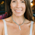 A smiling woman with long brown hair wearing a silver feather pendant necklace with a light blue beaded chain, paired with a white tank top.