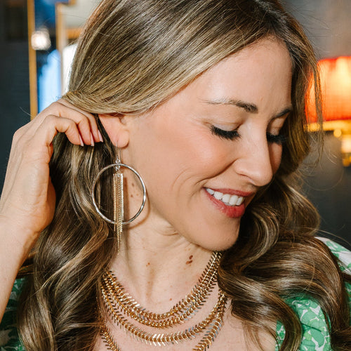 A woman with long wavy brown hair is shown in a close-up wearing large gold hoop earrings with dangling chain details, layered gold necklaces, and a green-and-white patterned top.